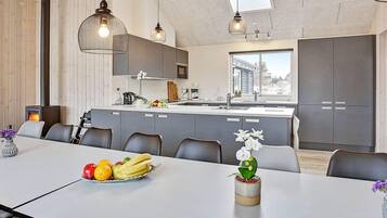Et moderne køkken med en central ø, hængende pendellamper og et stort vindue der lukker naturligt lys ind.
