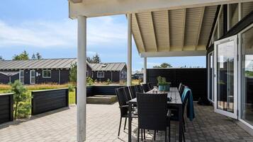 En udendørs patio med et bord og stole, en pergola over hovedet, og udsigt til boligbygninger i baggrunden.