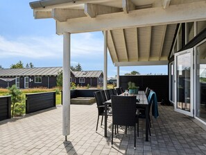 En udendørs patio med et bord og stole, en pergola over hovedet, og udsigt til boligbygninger i baggrunden.