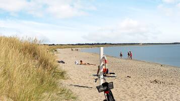 En strand med en cykel parkeret i sandet, en redningskrans hængende på en stang, og mennesker i det fjerne.