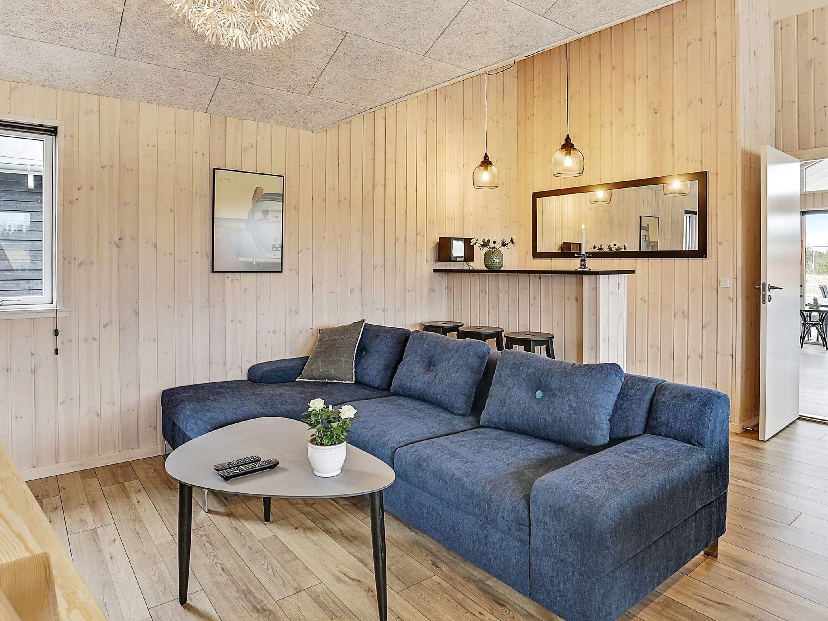 En moderne stue med en hjørnesofa, et sofabord og et lille sengebord. Et indrammet billede på væggen, et spejl over sengebordet, samt en loftlampe.