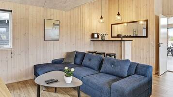 En moderne stue med en hjørnesofa, et sofabord og et lille sengebord. Et indrammet billede på væggen, et spejl over sengebordet, samt en loftlampe.