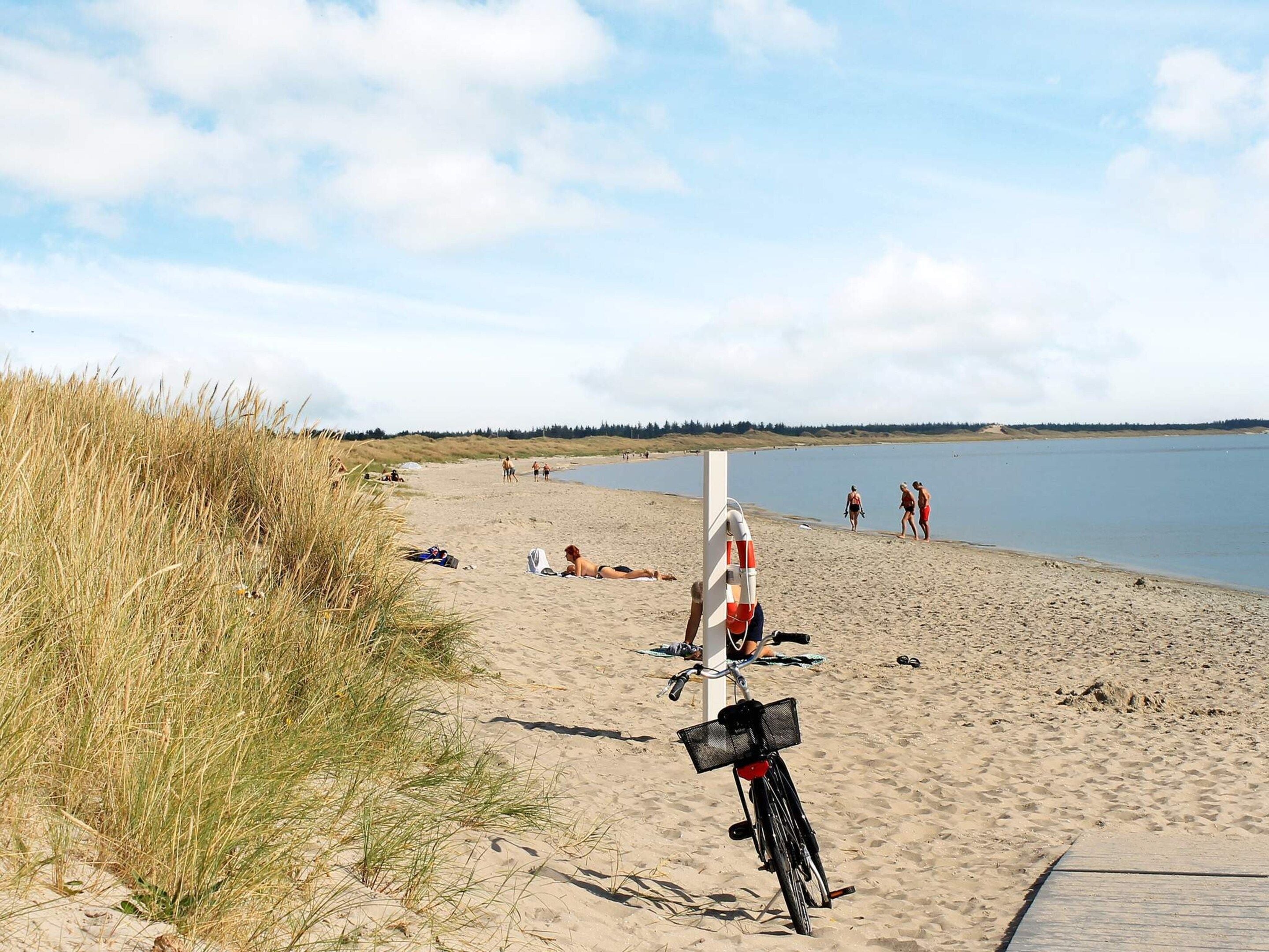 En strand med en cykel parkeret i sandet, en redningskrans fastgjort til en stang, og mennesker i baggrunden.
