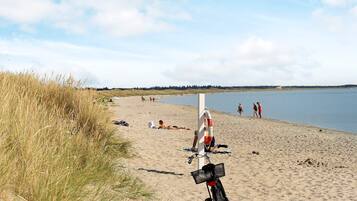 En strand med en cykel parkeret i sandet, en redningskrans fastgjort til en stang, og mennesker i baggrunden.