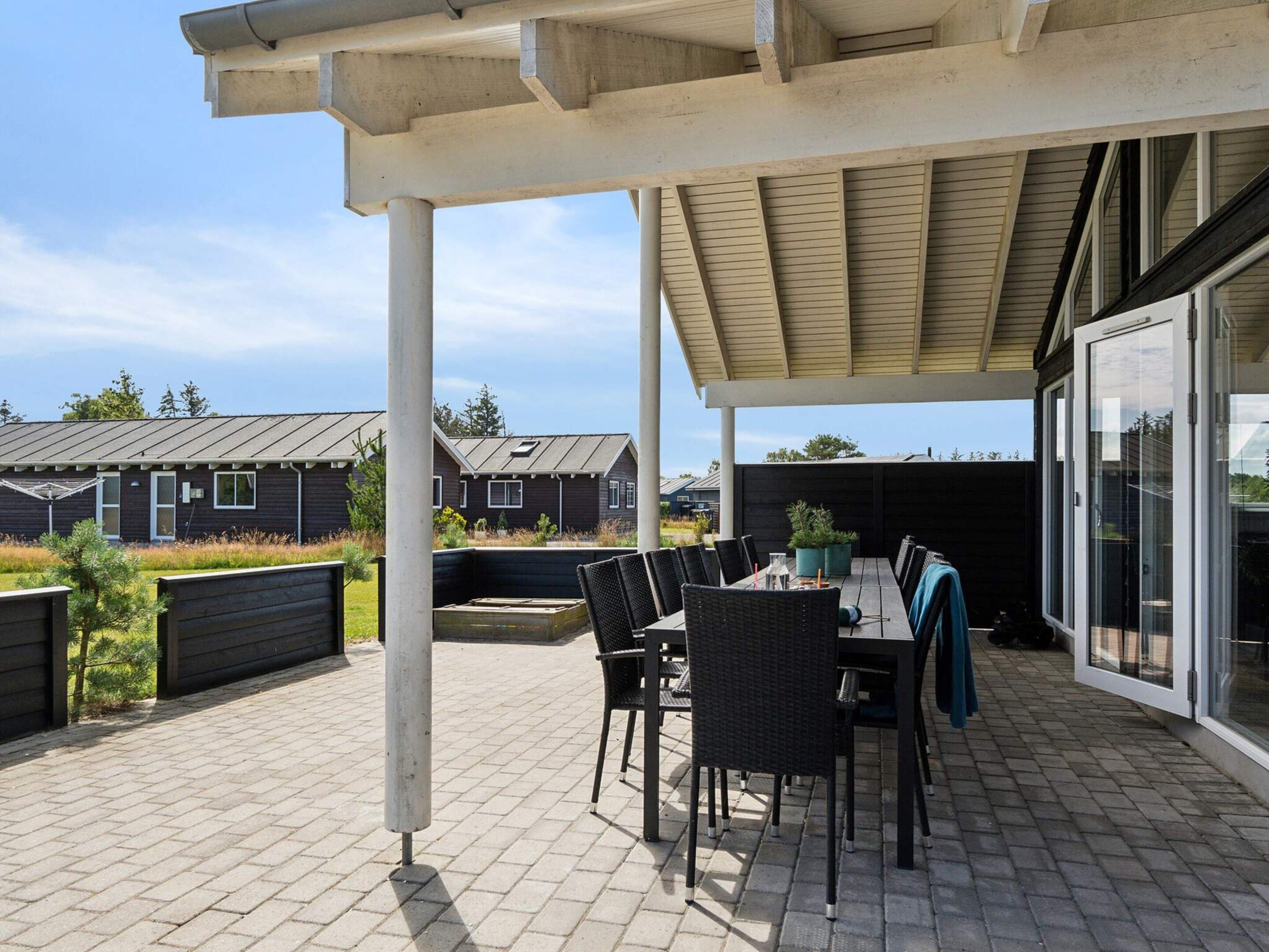 A covered outdoor patio with a table and chairs, a pergola overhead, and a view of residential buildings in the background.