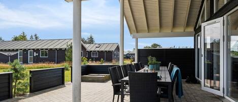A covered outdoor patio with a table and chairs, a pergola overhead, and a view of residential buildings in the background.