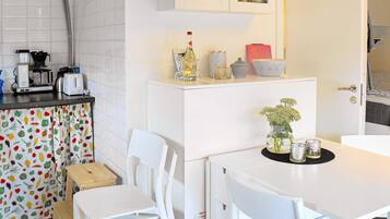 A compact kitchen with a white countertop, a coffee machine, a toaster, and a small refrigerator. There is a dining area with a white table and chairs.