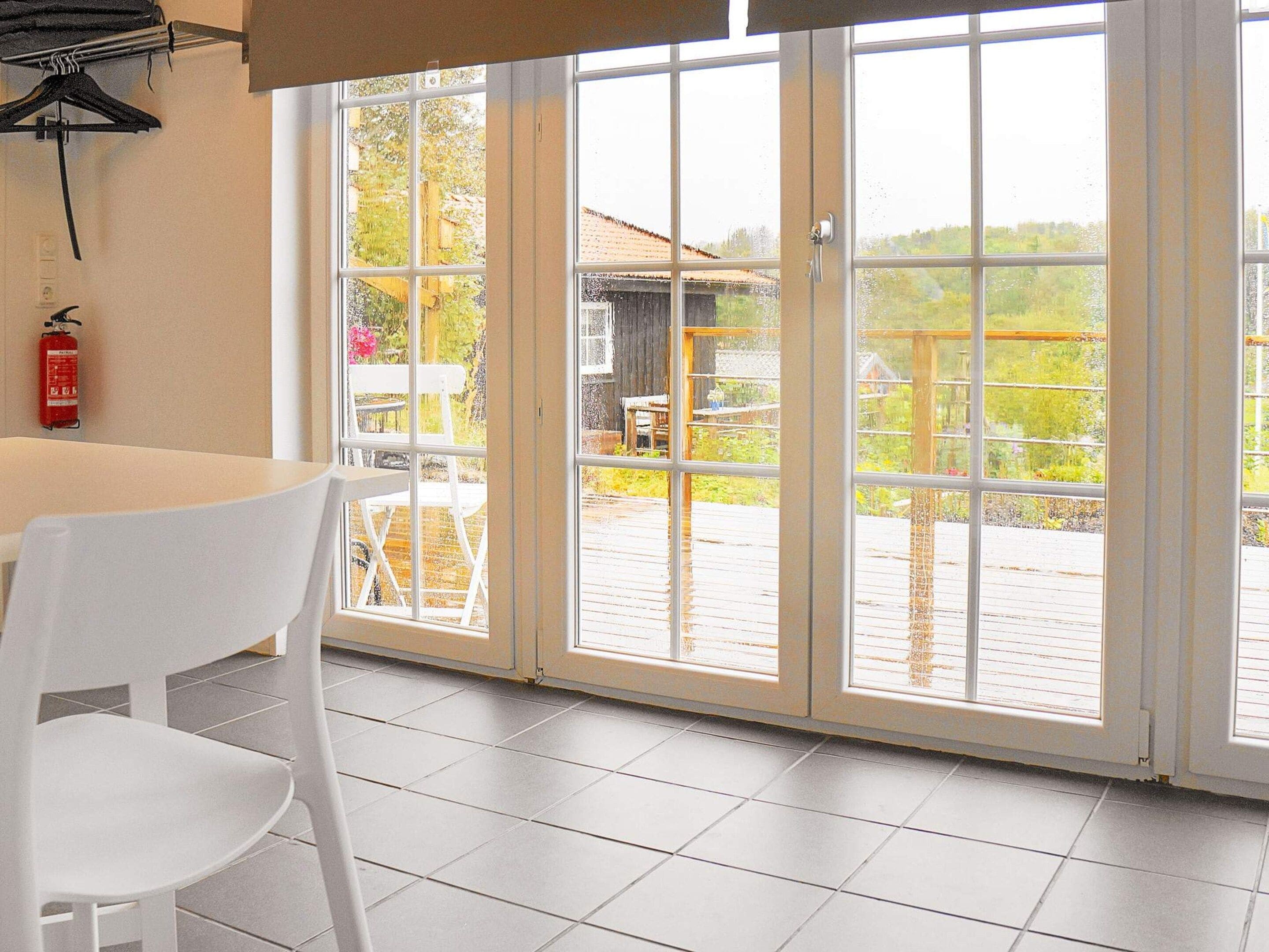 Una habitación con una mesa y silla blancos, un extintor en la pared y vistas a una terraza exterior a través de puertas de cristal.