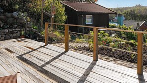 Um deck de madeira com guarda-corpo de metal, um pequeno edifício de madeira e um muro de pedra ao fundo.