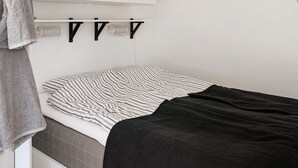 Quarto compacto com cama, toalha, armário branco e luminária fixa na parede.