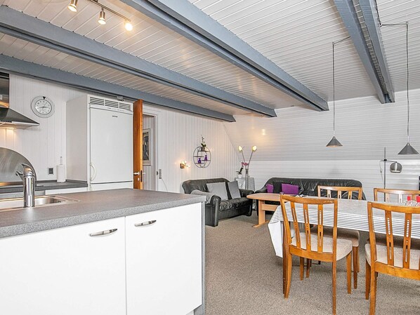A modern kitchen with a built-in oven, a dining area with wooden chairs and a table, and a living area with a sofa and decorative items.
