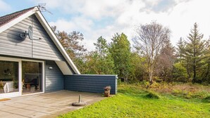 Une maison moderne avec un toit en pente, une terrasse avec une table et des chaises, et un jardin agrémenté d’un petit abri de jardin.