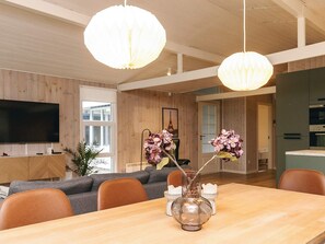 A modern living room with a wooden ceiling, a dining table with chairs, a TV mounted on the wall, and a vase with flowers on the table. - 8 Person Holiday Home in Hadsund-by Traum (Hadsund)
