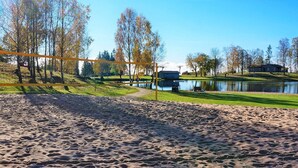 A sandy beach area with a volleyball net, a pond, and trees in the background.