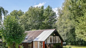 A small, single-story house with a sloped metal roof, large windows, and a wooden deck. - 4 Person Holiday Home in Oksbol (Oksbol)