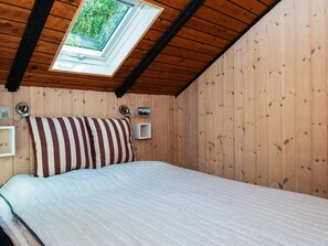 A bedroom with a wooden ceiling, a skylight, a bed with striped bedding, a nightstand, and a wall-mounted light.