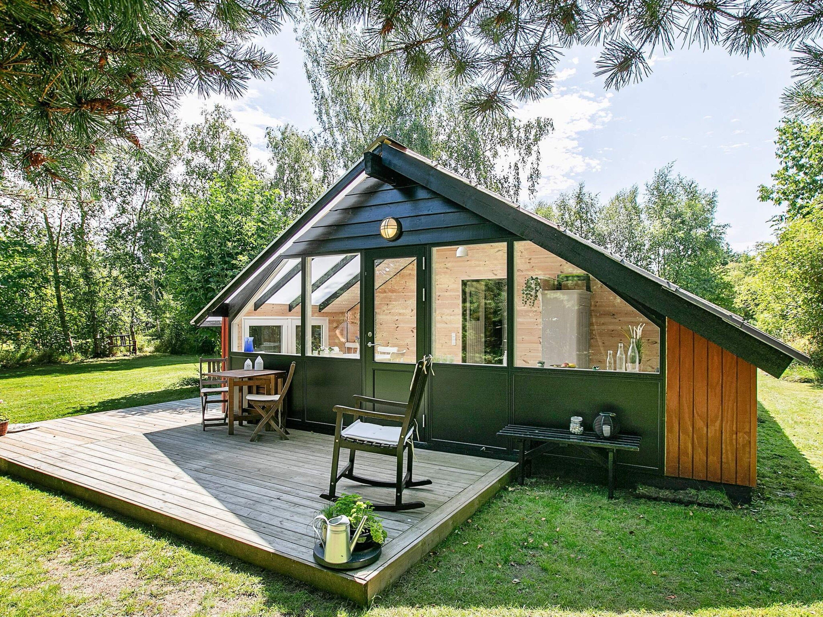 A modern wooden cabin with a deck, a dining area, and a large window overlooking a garden.