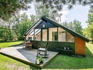 A modern wooden cabin with a deck, a dining area, and a large window overlooking a garden.
