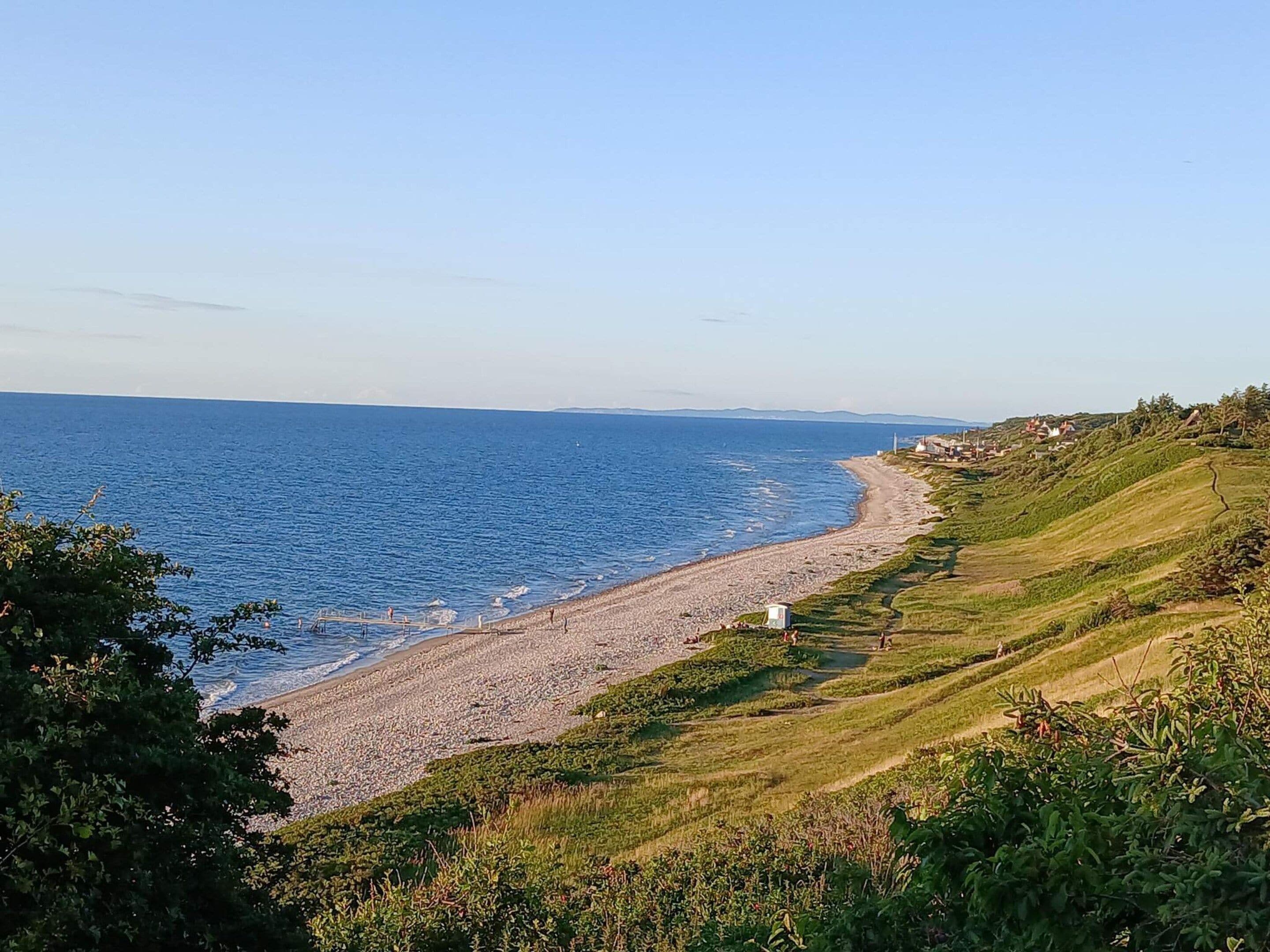 A coastal landscape with a pebble beach, grassy hills, and a clear sky.