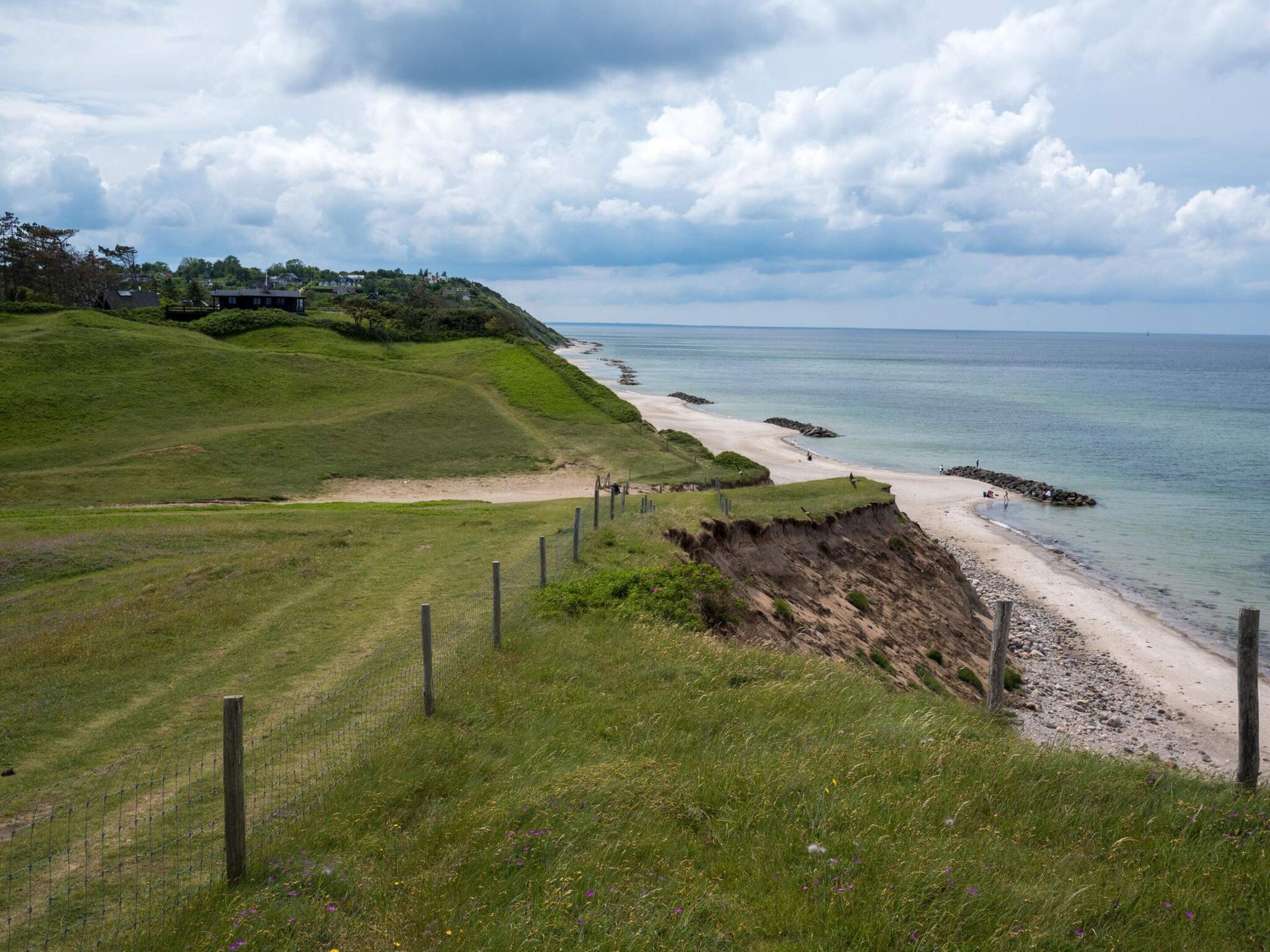 Un paysage côtier avec un sentier herbeux menant à une plage, une clôture et des maisons en arrière-plan.