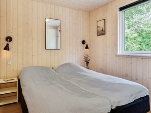A room with wooden panel walls, a bed with a striped bedspread, a bedside table with a book, a wall-mounted mirror, a picture, and a window with a view of greenery. - Chic Holiday Home in Hovedstaden near Sea (Vejby)