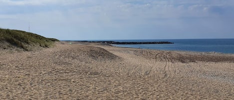Uma praia de areia com uma duna gramada e um afloramento rochoso ao longe.