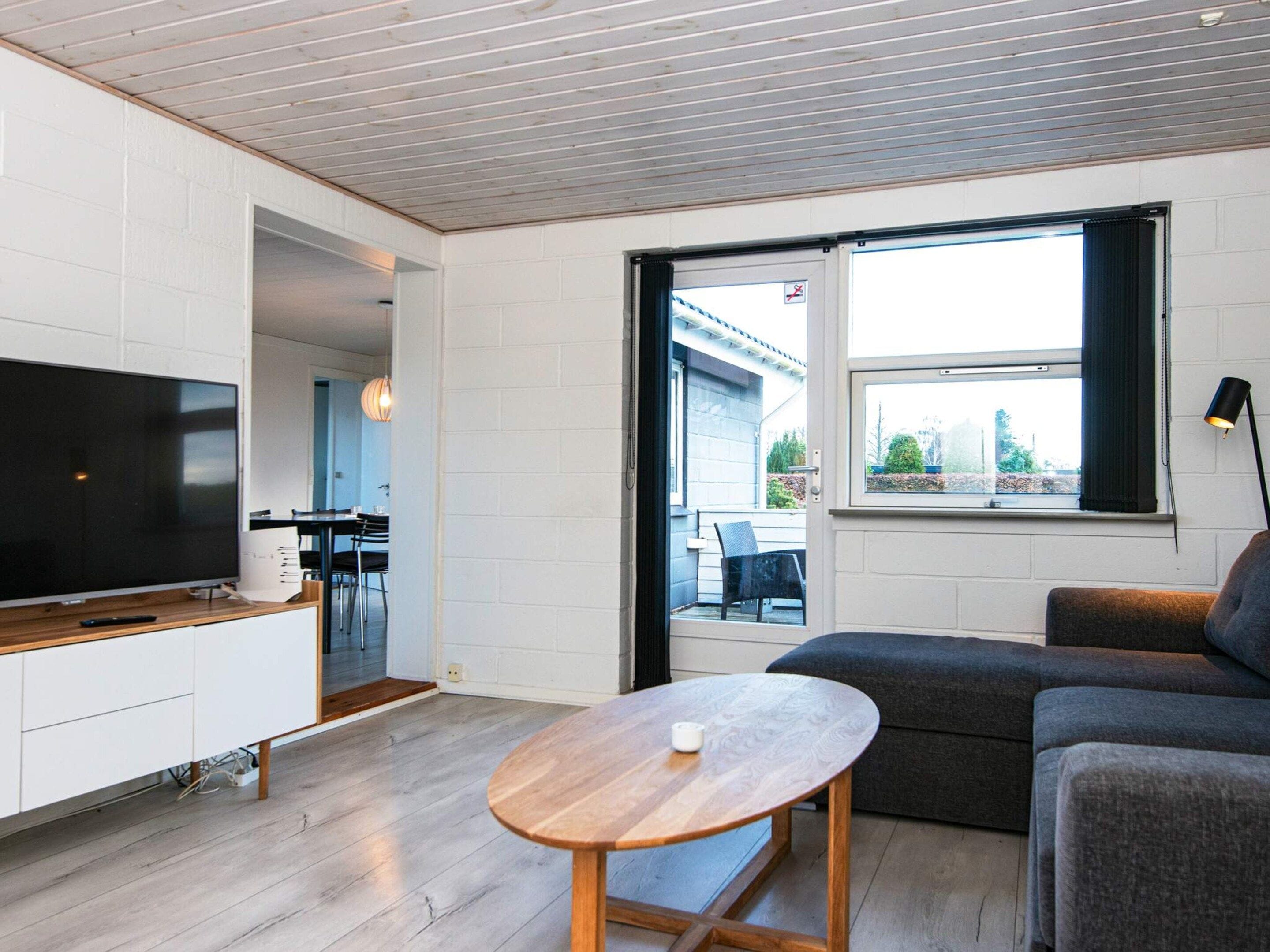 Una sala de estar moderna con una televisión de pantalla plana, una mesa redonda de madera, un sofá gris y una ventana con vistas al exterior.