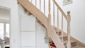 Una escalera de madera con barandilla, una pared blanca y una puerta que da a un cuarto con un armario blanco y una pequeña mesa con sillas.