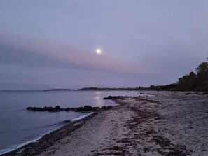 Una playa con costa rocosa, agua tranquila y una luna visible en el cielo.