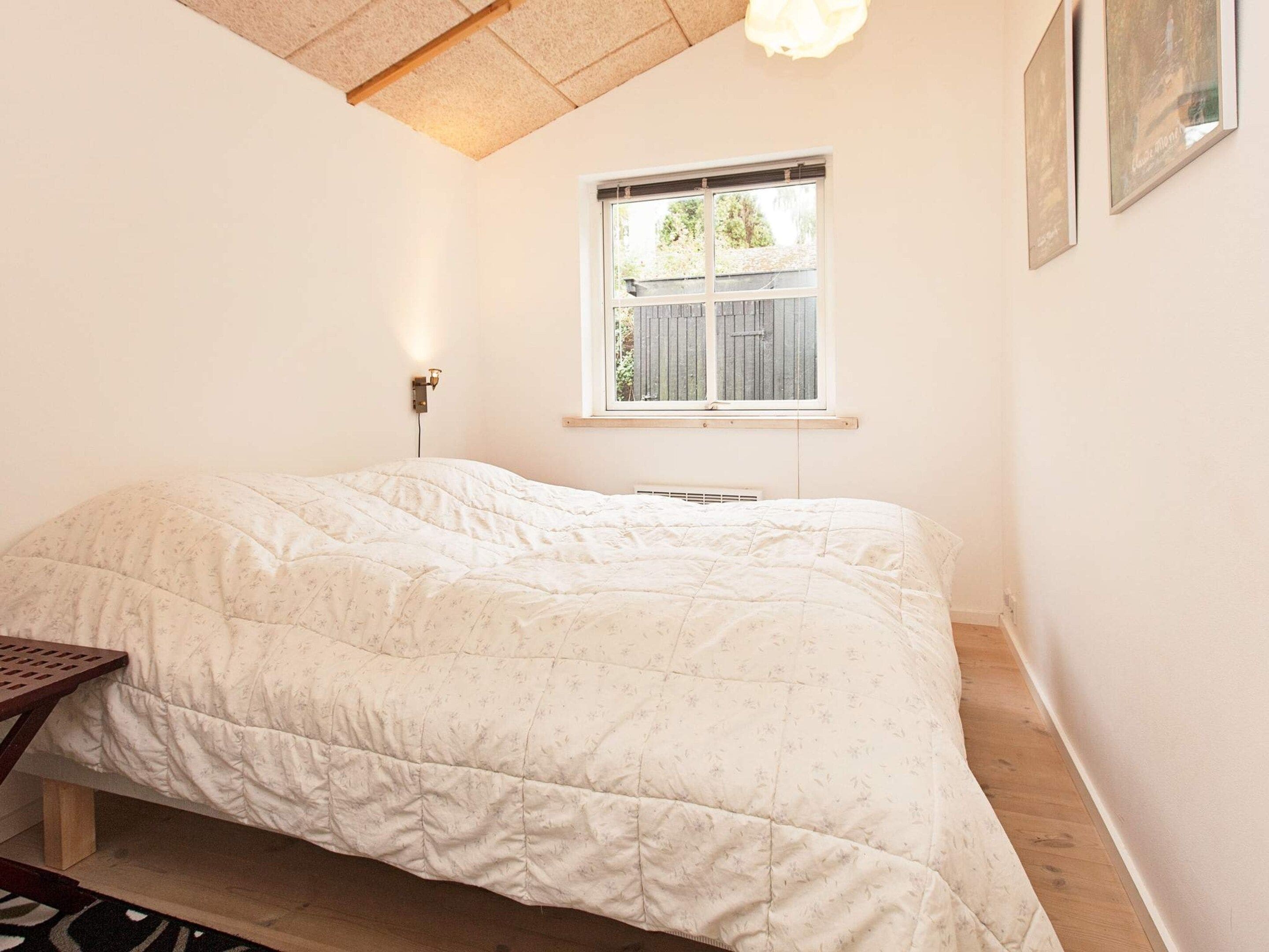 A bedroom with a bed, a window, a wall-mounted light, and a wooden floor.