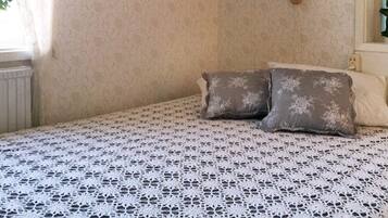 A room with a bed covered in a white lace bedspread, two pillows, a wall-mounted light fixture, and a radiator.