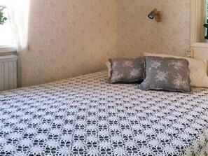 A room with a bed covered in a white lace bedspread, two pillows, a wall-mounted light fixture, and a radiator. - Holiday Home in Kode (Kode)