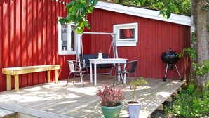 A wooden deck with a red building in the background, a table with chairs, a bench, and a grill.