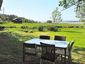 Mesa e quatro cadeiras em ambiente externo, com um campo gramado, um muro de pedra e um edifício de madeira ao fundo.