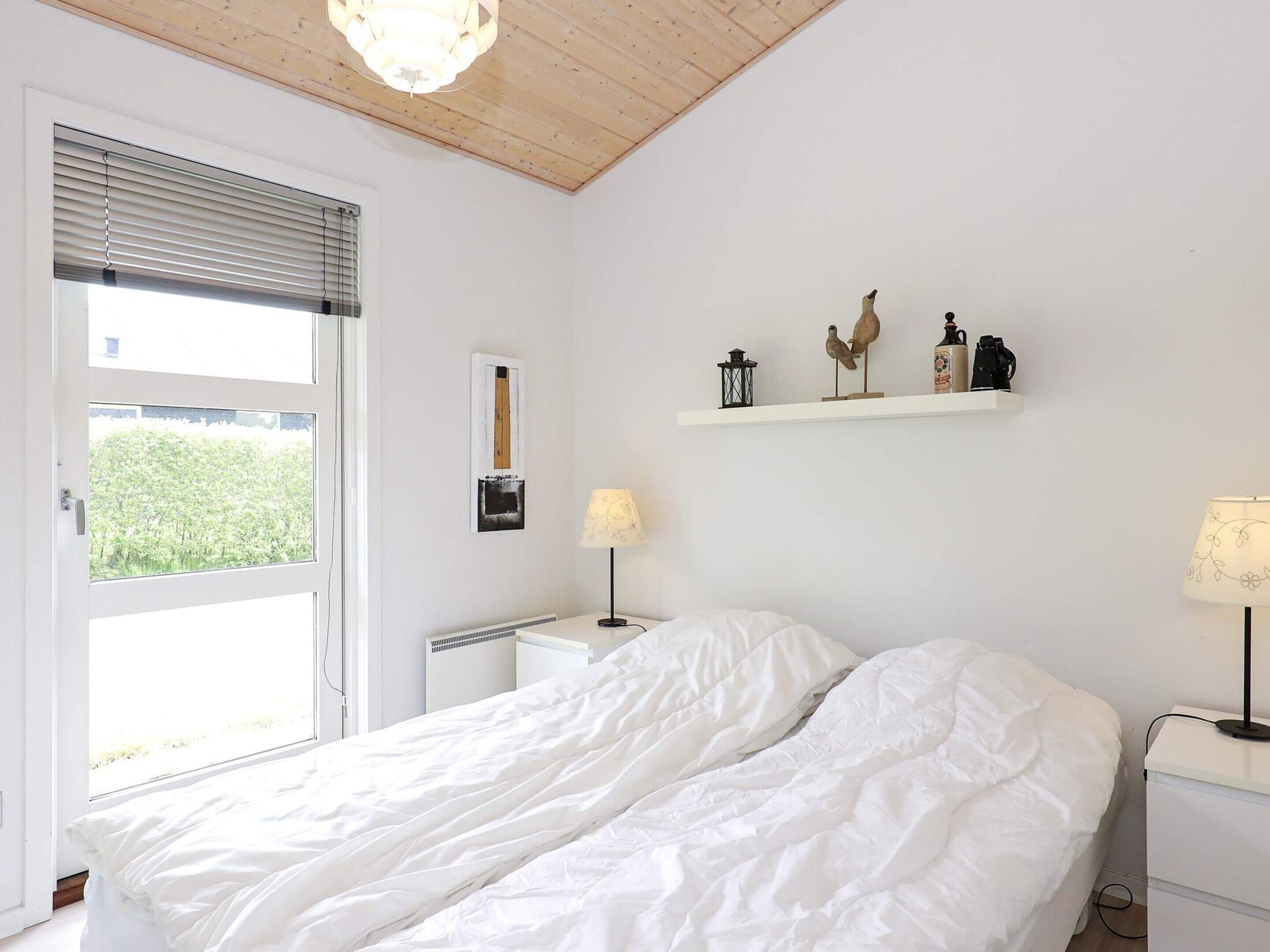 A bedroom with a bed, a nightstand, a lamp, a shelf with decorative items, and a window with blinds.