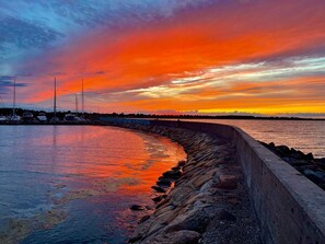 A waterfront scene with a curved stone wall, calm water reflecting the sunset, and boats docked in the background.