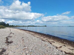 A pebble beach with calm water, distant land, and a partly cloudy sky.