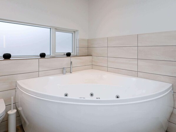 A modern bathroom with a large white bathtub, a toilet, and a window above the bathtub.