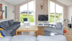 A spacious living room with a large TV, grey sofas, a wooden coffee table, and a view of the outdoors through large windows.