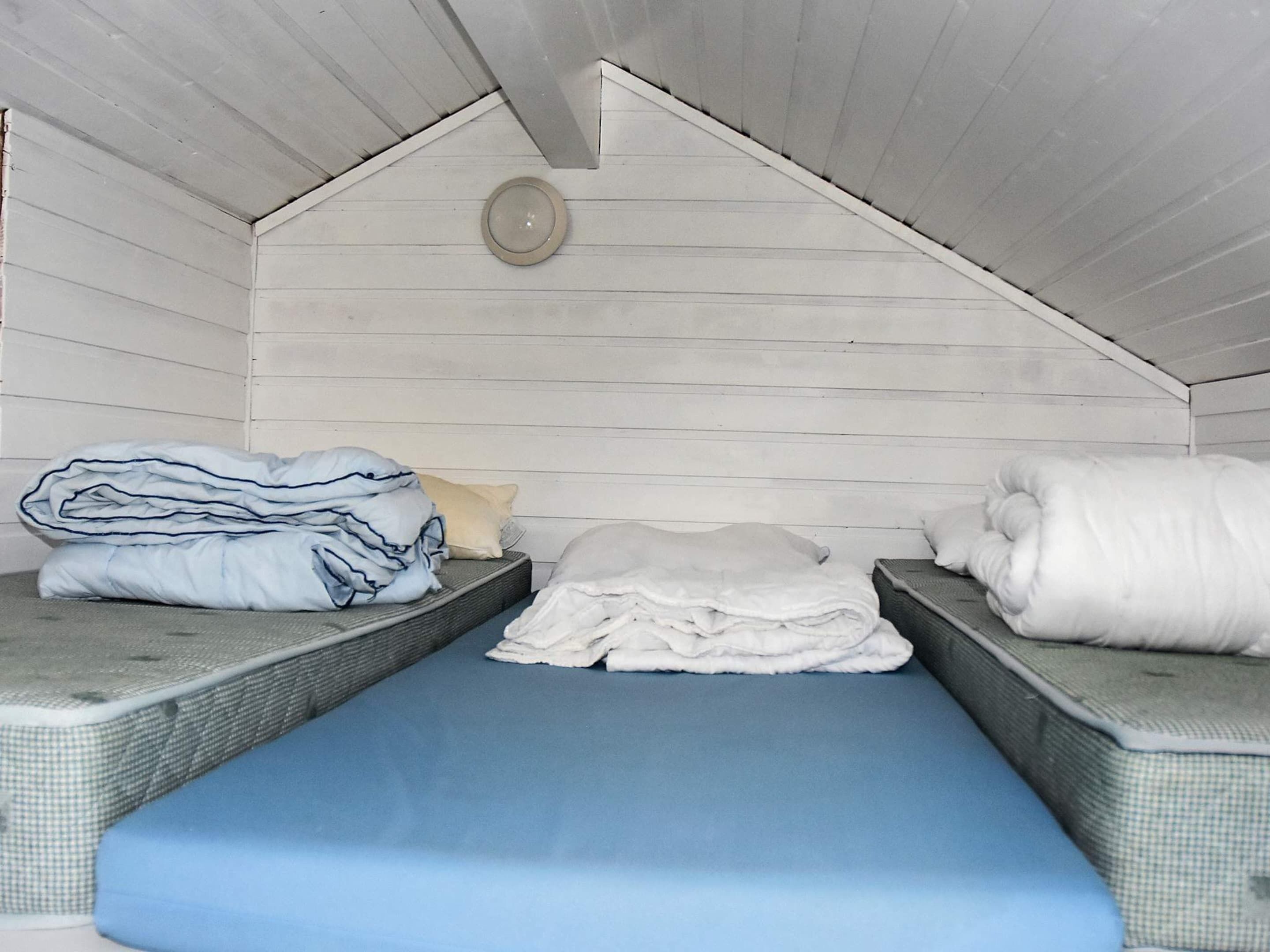 Une petite cabane d’une seule pièce, avec un plafond en pente, un lit au centre surmonté d’un matelas bleu, et deux bancs recouverts de couvertures pliées.