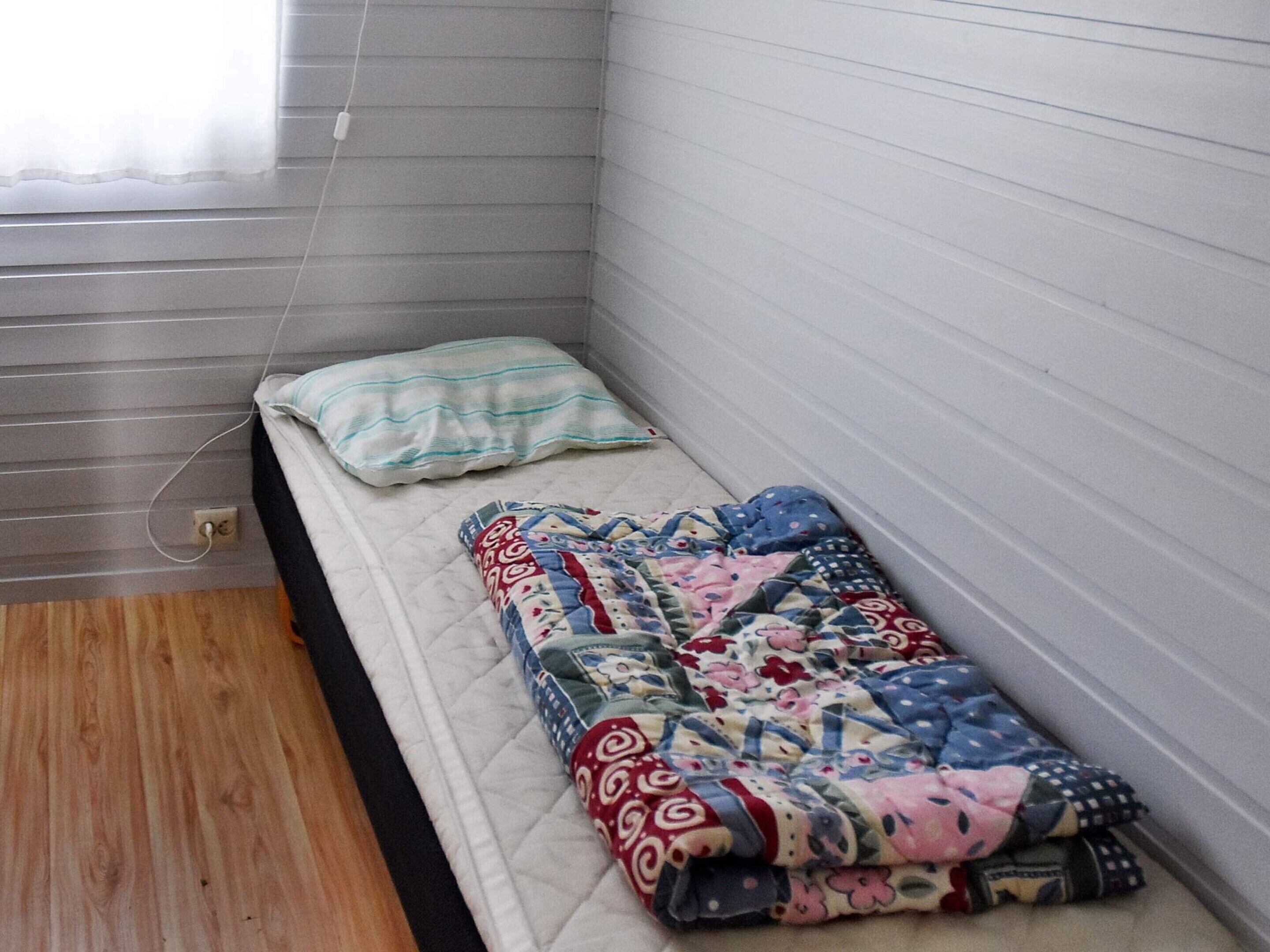 Une petite pièce étroite, avec un lit adossé au mur, une fenêtre avec des stores et une prise de courant au sol.