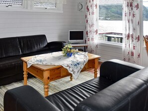 A living room with a black leather sofa, a wooden coffee table with a floral tablecloth, a television on a stand, and a window with curtains.