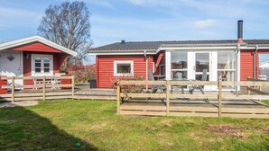 A red wooden house with a white door and windows, a chimney, and a fenced yard with a picnic table.