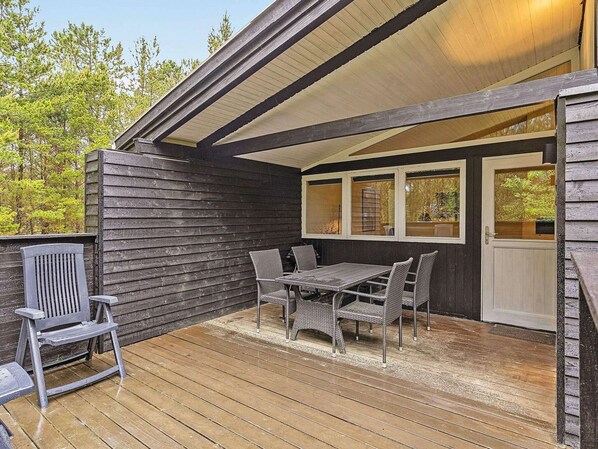 Une terrasse extérieure couverte, dotée d’une table et de chaises, d’une terrasse en bois et offrant une vue sur les arbres environnants.