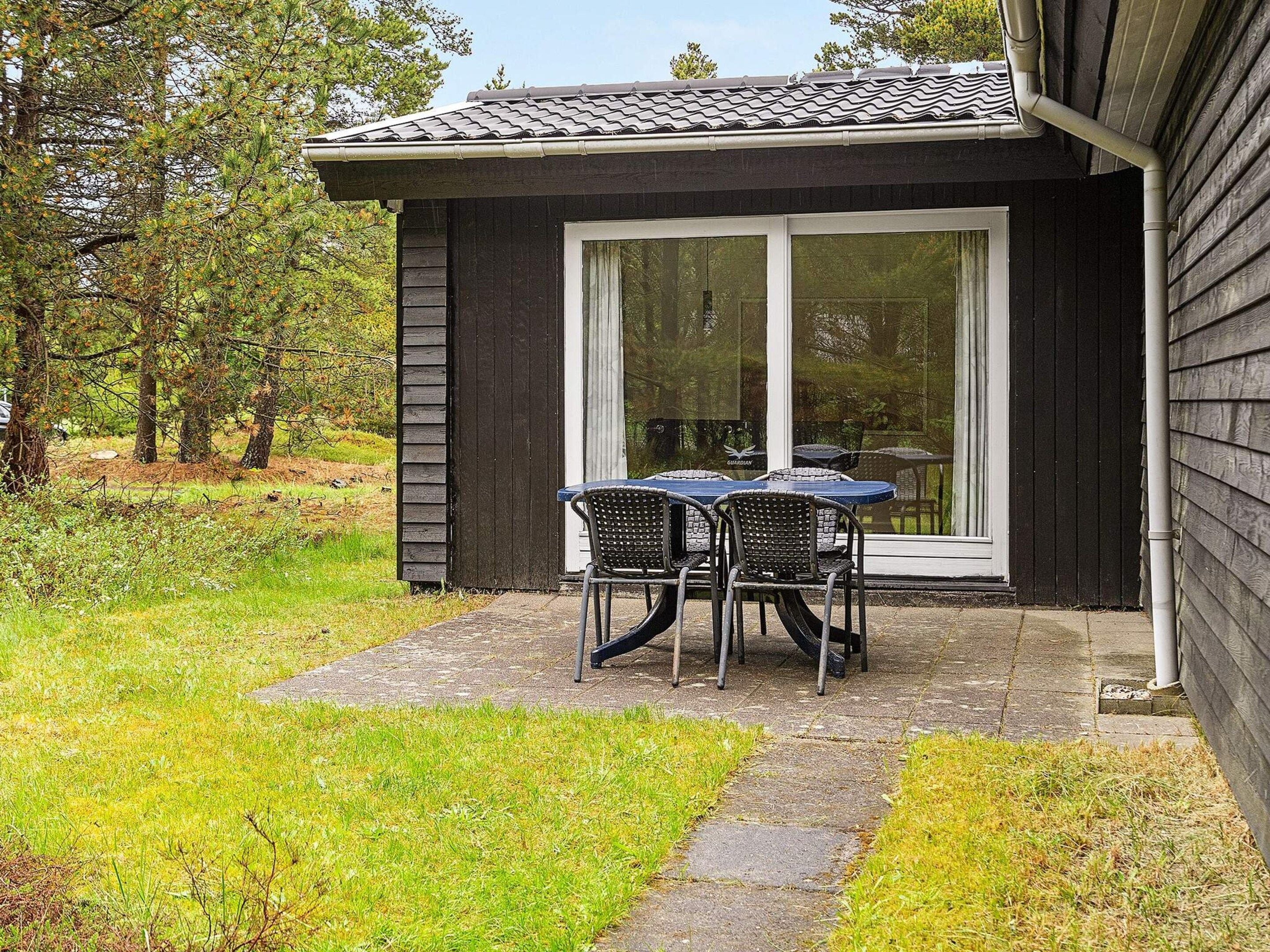 Een klein gebouw met een terras, inclusief een tafel en stoelen, omgeven door een gazon en bomen.