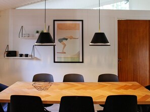 A dining area with a wooden herringbone table, black chairs, pendant lights, a framed picture, and a small shelf with items.