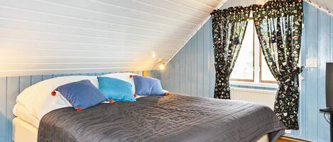 A bedroom with a sloped ceiling, a bed with multiple pillows, a window with floral curtains, and a wooden floor.