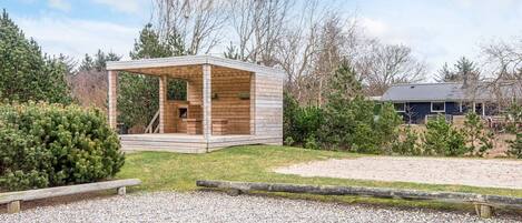Una cabaña de madera con porche cubierto, rodeada de grava y follaje, con una casa al fondo.