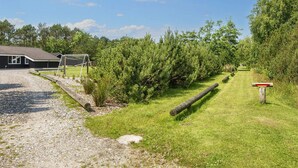 Un chemin de gravier mène à un bâtiment où se trouve un but de sport, le tout étant entouré de verdure et d’arbres.