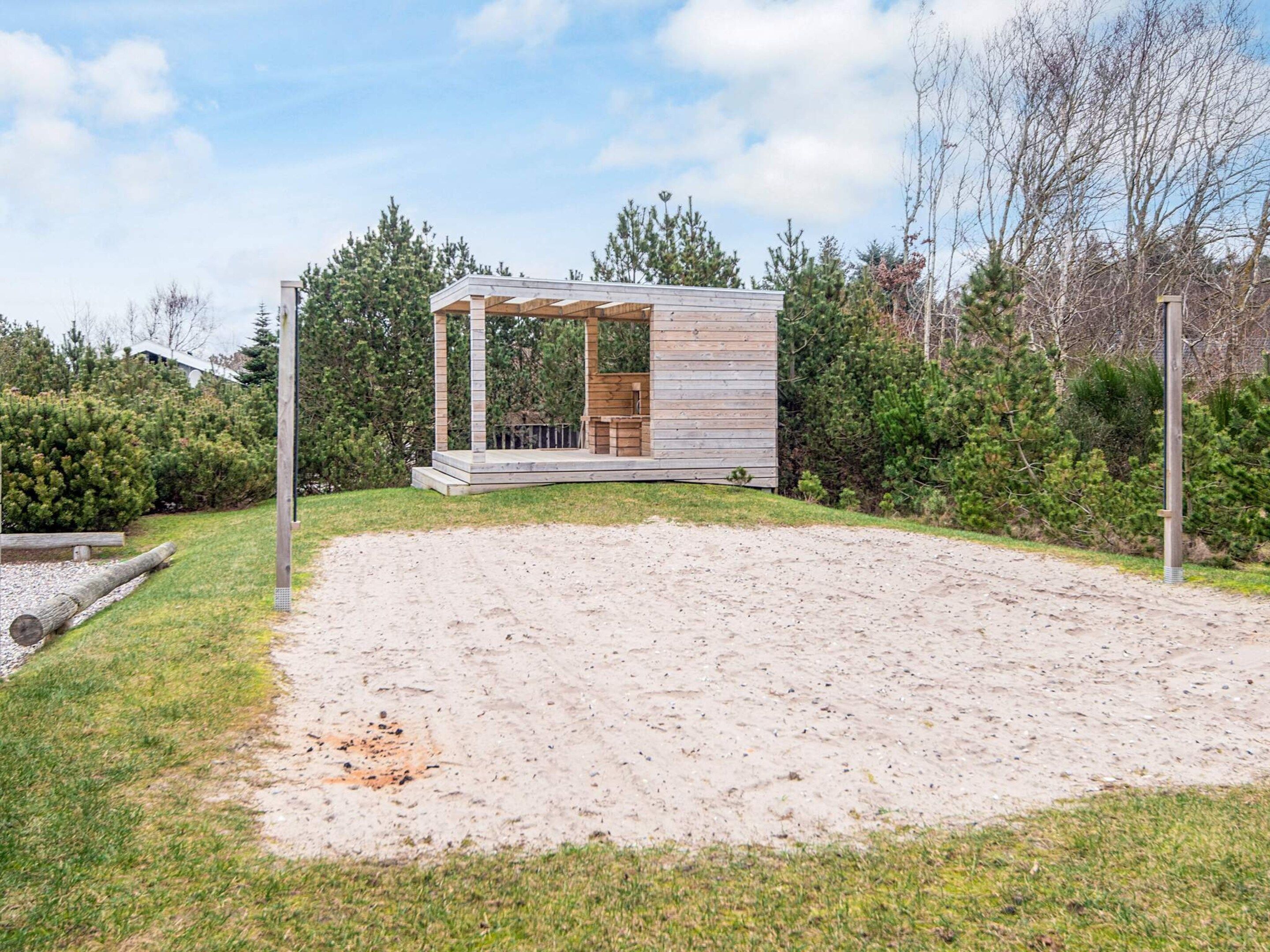 A wooden cabin with a covered porch, surrounded by a sandy area and dense greenery.
